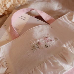 May include: A white tote bag with a pink handle and ruffled edges. The bag features an embroidered design of a rabbit and flowers. Books are visible inside the bag. The bag is made of a soft, textured fabric.
