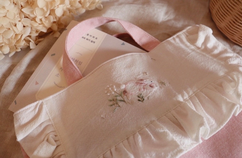 May include: A white tote bag with a pink handle and ruffled edges. The bag features an embroidered design of a rabbit and flowers. Books are visible inside the bag. The bag is made of a soft, textured fabric.