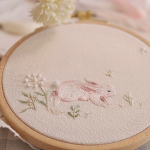May include: Embroidered artwork in a wooden hoop. The design features a pink and white bunny lying in a field of green and white embroidered flowers and foliage. The background is a light beige fabric.