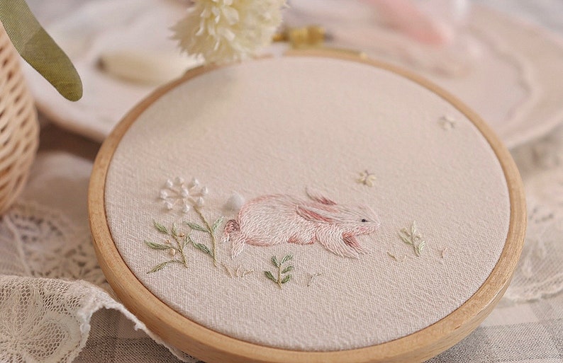 May include: Embroidered artwork in a wooden hoop. The design features a pink and white bunny lying in a field of green and white embroidered flowers and foliage. The background is a light beige fabric.