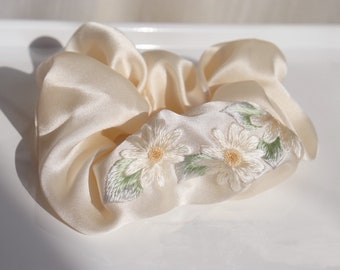 Puede incluir: Un scrunchie de satén crema con margaritas blancas y hojas verdes bordadas.