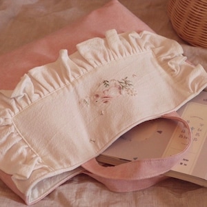 May include: A pink tote bag with a white eye mask featuring floral embroidery and ruffled edges. A book with Korean text is inside the bag. The scene is set on a pink surface, with a wicker basket in the background.