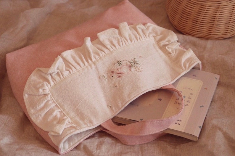 May include: A pink tote bag with a white eye mask featuring floral embroidery and ruffled edges. A book with Korean text is inside the bag. The scene is set on a pink surface, with a wicker basket in the background.