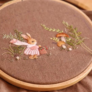 May include: Embroidered hoop art featuring a brown rabbit wearing a pink dress and carrying a basket of flowers. The rabbit is surrounded by green foliage and brown mushrooms.