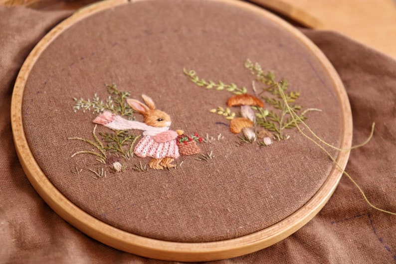 May include: Embroidered hoop art featuring a brown rabbit wearing a pink dress and carrying a basket of flowers. The rabbit is surrounded by green foliage and brown mushrooms.