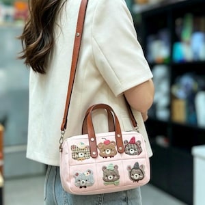 Puede incluir: Un bolso rosa con asa y correa de cuero marrón. El bolso presenta seis caras de oso bordadas con varios accesorios. El bolso es rectangular y tiene cierre de cremallera.