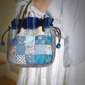 May include: A patchwork bag with a variety of blue fabric squares, including floral, polka dot, and striped patterns. The bag has brown leather handles and a drawstring closure. The bottom edge is trimmed with a gray and white herringbone pattern.