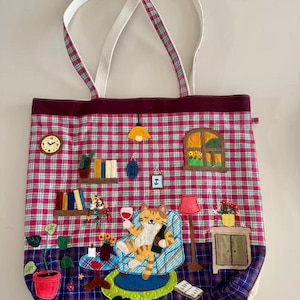 May include: A whimsical tote bag with a plaid pattern in pink, white, and burgundy. The bag features an appliqued scene of a cat relaxing in a chair, holding a wine glass and bottle. Additional appliqued details include a clock, books, and a lamp.