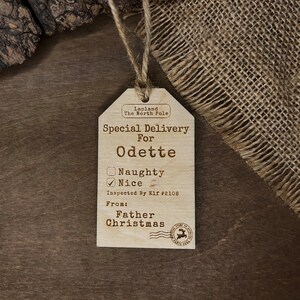 May include: A wooden Christmas gift tag with the text "Lapland The North Pole" and "Special Delivery For Odette". The tag has a checkbox for "Naughty" and "Nice", with "Nice" checked. The tag also includes the text "Inspected By Elf #2108" and "From: Father Christmas".