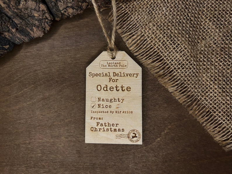 May include: A wooden Christmas gift tag with the text "Lapland The North Pole" and "Special Delivery For Odette". The tag has a checkbox for "Naughty" and "Nice", with "Nice" checked. The tag also includes the text "Inspected By Elf #2108" and "From: Father Christmas".