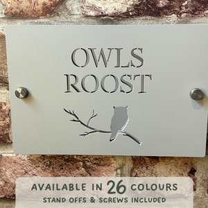 May include: A gray metal house number sign with the text "OWLS ROOST" and a cutout silhouette of an owl perched on a branch. The sign is mounted on a brick wall with screws. The text "AVAILABLE IN 26 COLORS" and "STAND OFFS & SCREWS INCLUDED" is visible below the sign.