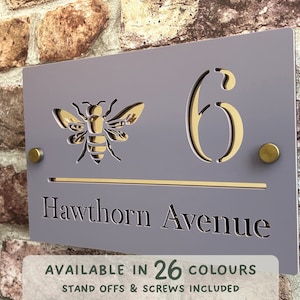 Può includere: Un cartello per numeri civici viola con un'ape dorata ritagliata e il numero 6. Il cartello dice anche "Hawthorn Avenue" in lettere dorate. Il cartello è disponibile in 26 colori.