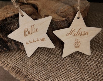May include: Two wooden star-shaped ornaments with twine hangers. One ornament is engraved with the name "Billie" and a reindeer and sleigh design. The other ornament is engraved with the name "Melissa" and a penguin design.