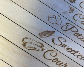Può includere: Un tagliere di legno con etichette per diverse verdure, tra cui 'Cavolo rosso', 'Patate', 'Mais', 'Zucchine', 'Piselli' e 'Fagioli'.