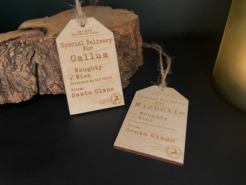 May include: Two wooden Christmas gift tags with a rustic design. The tags are brown and have a white string attached. The tags have the text "Special Delivery" and "Inspected By Elf #2108" on them. One tag is for Callum and the other is for Michelle. Both tags have a checkbox for "Naughty" and "Nice" with the "Nice" box checked.
