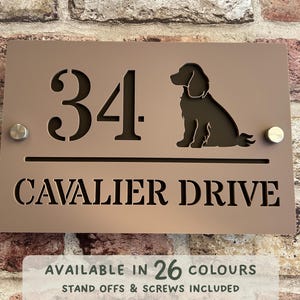 Op de afbeelding: Een bruin metalen huisnummerbord met het nummer 34 en de tekst "CAVALIER DRIVE" in een serif-lettertype. Boven de tekst is een silhouet van een hond uitgesneden. Het bord is verkrijgbaar in 26 kleuren.