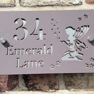 Può includere: Un cartello per numeri civici di colore lilla chiaro con il numero 34, le parole "Emerald Lane" e un disegno ritagliato di una fata con ali e fiori.