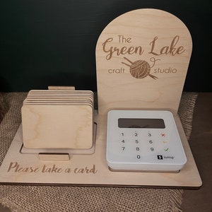 May include: A wooden stand with a sign that reads "The Green Lake craft studio" and a ball of yarn with knitting needles. The stand also has a card holder and a white credit card reader with a keypad and a screen that says "SumUp". The text "Please take a card" is engraved on the stand.