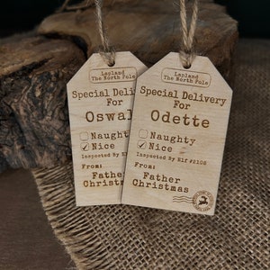May include: Two wooden Christmas gift tags with a rustic brown finish. Each tag has a checklist for "naughty" or "nice" and a space for a name. The tags read "Special Delivery for Oswald" and "Special Delivery for Odette". Both tags are labelled "Lapland The North Pole" and "From: Father Christmas".