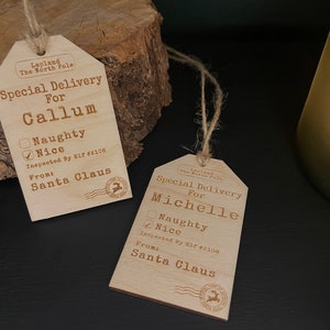 May include: Two wooden Christmas gift tags with a rustic design. The tags are shaped like a luggage tag and have a string attached. The tags are personalised with the names "Callum" and "Michelle" and have a checkbox for "Naughty" or "Nice". The tags also include the text "Special Delivery For" and "Inspected By Elf #2106" and "From: Santa Claus".