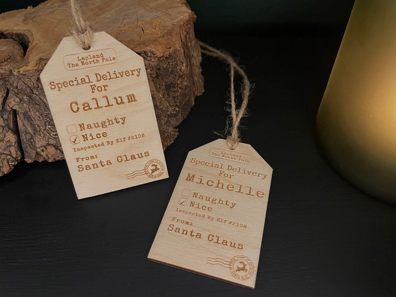 May include: Two wooden Christmas gift tags with a rustic design. The tags are shaped like a luggage tag and have a string attached. The tags are personalised with the names "Callum" and "Michelle" and have a checkbox for "Naughty" or "Nice". The tags also include the text "Special Delivery For" and "Inspected By Elf #2106" and "From: Santa Claus".