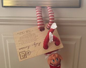 May include: A Christmas elf doll hanging upside down from a mailbox, holding a wooden postcard with the text "Molly & Harrison Greetings From the North Pole". The postcard has a red and white striped border and a red and white striped hat.