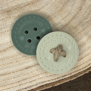May include: Two hand-knitted buttons in a light green colour. One button has a heart and the text "HANDKNITTED BY MELANIE". The other button has five holes and the text "HANDKNITTED BY ME".