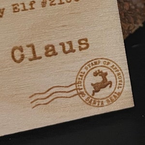 May include: A wooden tag with the text "Santa Claus" and a red reindeer stamp with the text "Official Stamps of Approval Santa Seal".