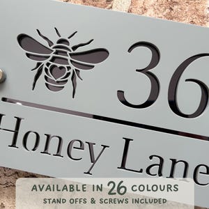 Può includere: Un cartello con il numero civico grigio chiaro con un disegno a forma di ape ritagliata e il numero 36. Il cartello recita "Honey Lane" e include il testo "Disponibile in 26 colori". Include distanziatori e viti.