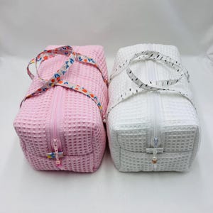 Può includere: Due borse da viaggio quadrate, una rosa e una bianca. La borsa rosa ha un cinturino floreale e quella bianca un cinturino a pois neri. Entrambe hanno una trama a nido d'ape, chiusura con zip e un ciondolo a perla. Ideali per articoli da toeletta.