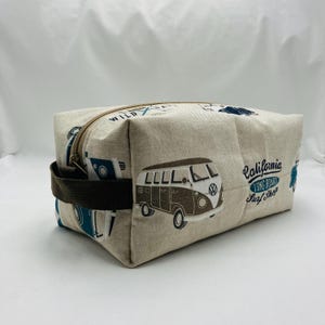 Op de afbeelding: Beige rechthoekige reistas met een bruine rits en handvat. De tas heeft een herhalend patroon van blauwe en bruine surfbusjes en de woorden "California Long Board Surf Shop".