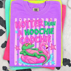 Puede incluir: Camisetas apiladas en tonos morado, rosa, verde y amarillo. La camiseta superior es morada con el texto "Hotter Than a Hoochie Coochie" y un gr&aacute;fico de un caim&aacute;n y un flamenco en un flotador inflable.