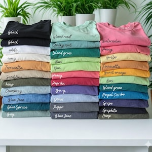 May include: Stacks of folded t-shirts in various colors, including black, white, and blue. Each stack is labeled with the color name in handwritten script. The shirts are arranged on a white surface with green plants in the background.