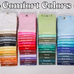 Puede incluir: Una exhibici&oacute;n de camisetas unisex Comfort Colors dobladas en varios colores, incluyendo blanco, rosa, verde y azul. Cada pila est&aacute; etiquetada con el nombre del color, como "Blossom" y "True Navy". El texto "Comfort Colors" se muestra en la parte superior.