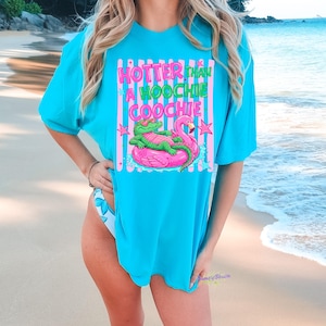 Puede incluir: Una camiseta turquesa con la frase "HOTTER than A HOOCHIE COOCHIE" en rosa y verde. El gr&aacute;fico presenta un caim&aacute;n con gafas de sol en un flotador de flamenco rosa. La camiseta se usa en la playa.