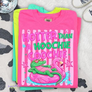 Puede incluir: Una pila de tres camisetas de colores ne&oacute;n: rosa, verde y amarillo. La camiseta rosa superior presenta el texto "HOTTER THAN A HOOCHIE COOCHIE" con un cocodrilo y un flamenco de dibujos animados en un flotador inflable. Las camisetas est&aacute;n dobladas.