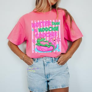 Puede incluir: Camiseta rosa brillante con la frase "Hotter Than A Hoochie Coochie" en verde y rosa. El gr&aacute;fico muestra un caim&aacute;n verde con gafas de sol en un flotador de flamenco rosa. Estilo casual.