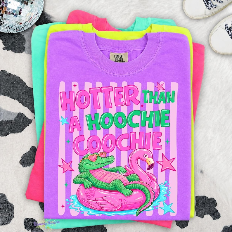 Puede incluir: Camisetas apiladas en tonos morado, rosa, verde y amarillo. La camiseta superior es morada con el texto "Hotter Than a Hoochie Coochie" y un gr&aacute;fico de un caim&aacute;n y un flamenco en un flotador inflable.