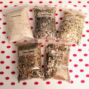 May include: Five clear plastic bags filled with various seed and grain mixes. Each bag has a label with a different name: "Calcium Combo", "A Little Seedy", "Coconut Chicken", "Poppin' Protein", and "Fall Harvest". The bags are arranged on a white surface with red polka dots.