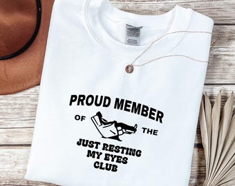 May include: White crewneck sweatshirt with the text "PROUD MEMBER OF THE JUST RESTING MY EYES CLUB" in black. A brown hat and a gold necklace are visible in the background. The sweatshirt is on a wooden surface.