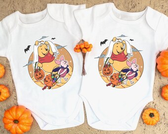 May include: Two white short-sleeve baby bodysuits featuring a graphic print of Winnie the Pooh wearing a ghost sheet and Piglet in a vampire cape, both holding jack-o'-lantern candy buckets against a circular tan background with bats.