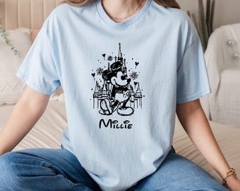 May include: Light blue t-shirt featuring a black and white graphic of Mickey Mouse in front of a castle with fireworks. The name "Millie" is printed below the graphic. The shirt is made of a soft material.