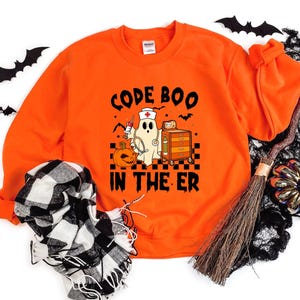 May include: A bright orange long-sleeve sweatshirt featuring a graphic print of a ghost dressed as a nurse with a syringe, a pumpkin, and a medical cart. The text reads CODE BOO IN THE ER in black letters above and below the central illustration.