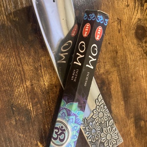 May include: A silver incense burner with a floral design and a box of HEM incense sticks with the word "OM" and a blue and purple mandala design. The box also has the word "HEM" printed on it.