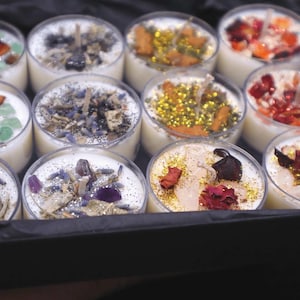 May include: Assortment of small, round candles in clear glass containers. Each candle has a different combination of dried flowers, herbs, and crystals embedded in the wax. The candles are arranged in a black box.