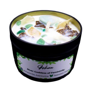 May include: A black candle tin with a white label that reads "Jshun River Goddess of Femininity Amazonite, Aventurine Seaweed, Seashells". The candle is topped with white wax and has green and brown crystals embedded in it.