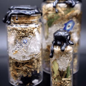 May include: Three glass jars with black wax seals, filled with dried herbs, crystals, and a silver charm with a blue eye. The jars are on a dark background.