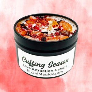 May include: A black metal tin candle with a white label reading Cuffing Season Love Attraction Candle BlkGrlMagick.com. The top of the candle features white wax embedded with orange tumbled stones, red dried flower petals, and a single central wick.