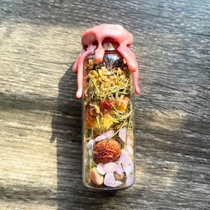 May include: A clear glass bottle filled with dried herbs, flowers, and pink crystals, sealed with a pink wax top.