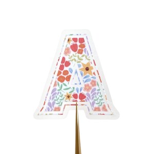Könnte beinhalten: Ein floral gemusterter Buchstabe "A" mit weißem Rand. Der Buchstabe ist mit einem farbenfrohen floralen Design mit roten, orangen, gelben und lila Blumen und grünen Blättern gefüllt. Der Buchstabe wird von einem goldfarbenen Stiel gehalten.